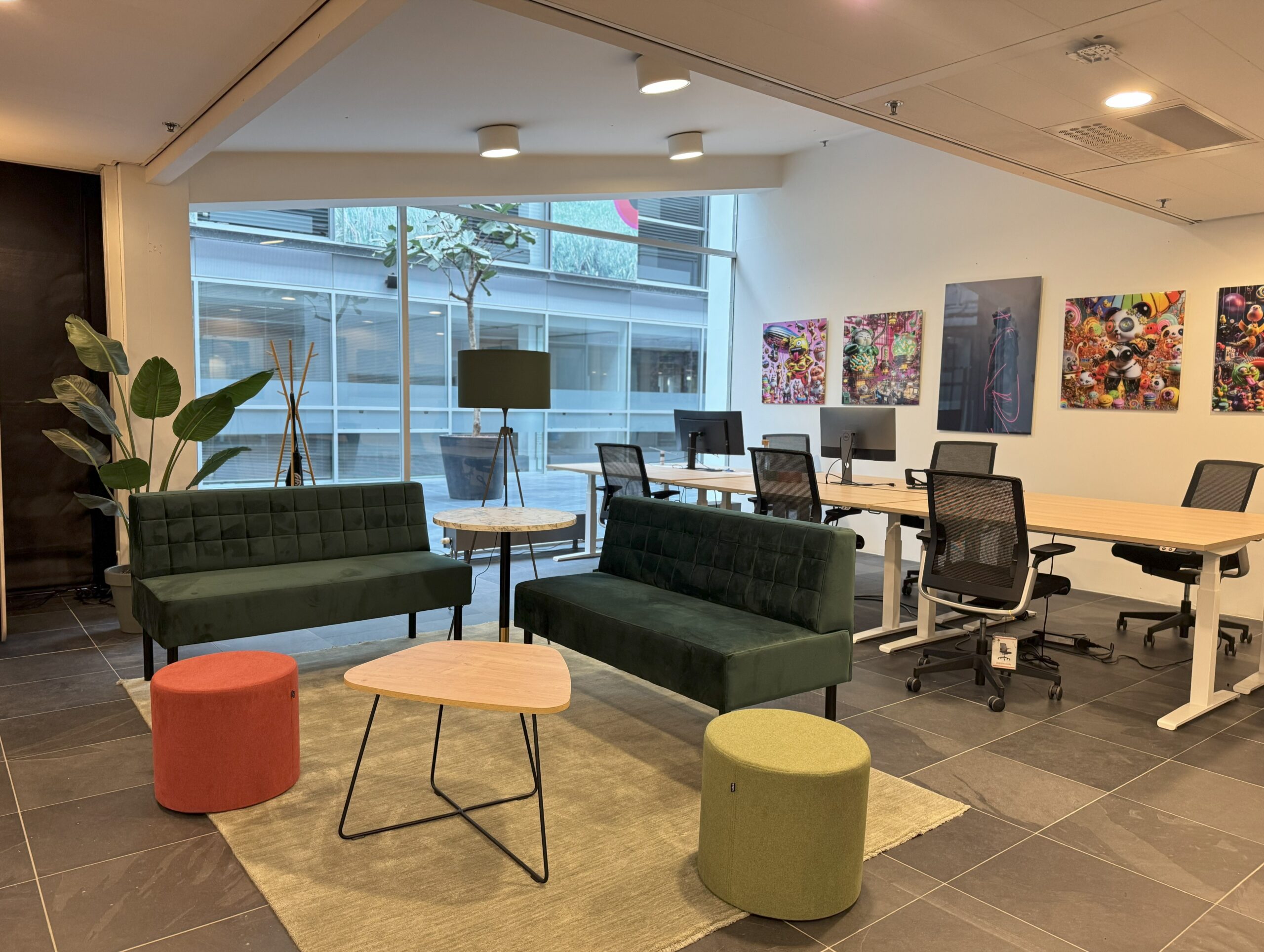 Moderne kantoorruimte met groene banken, kleurrijke kunst aan de muur en werkplekken met bureaus en stoelen.